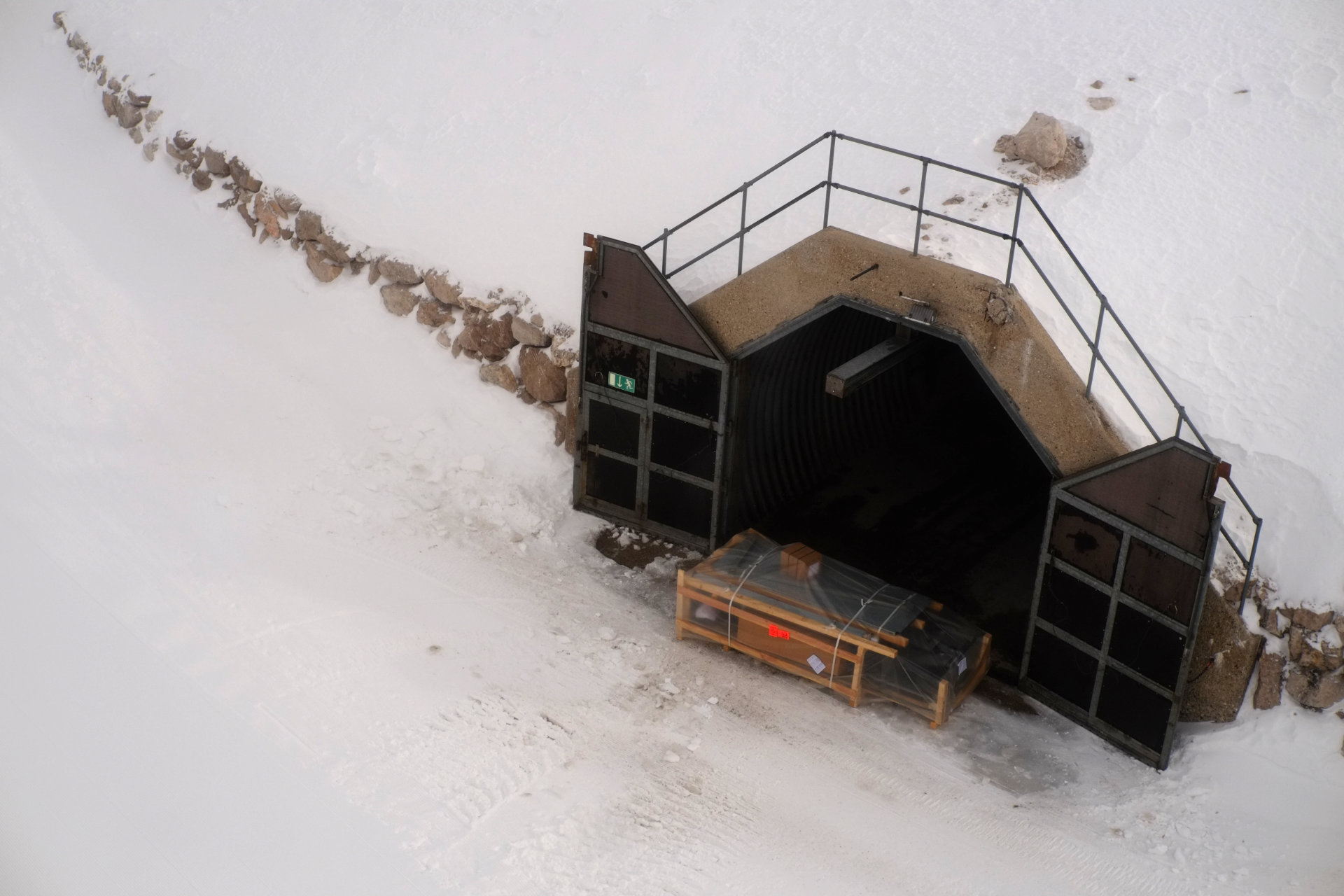 A photo of a mountain bunker tunnel with open doors, taken from above. A big wooden package wrapped in plastic lies in front of the tunnel entrance.