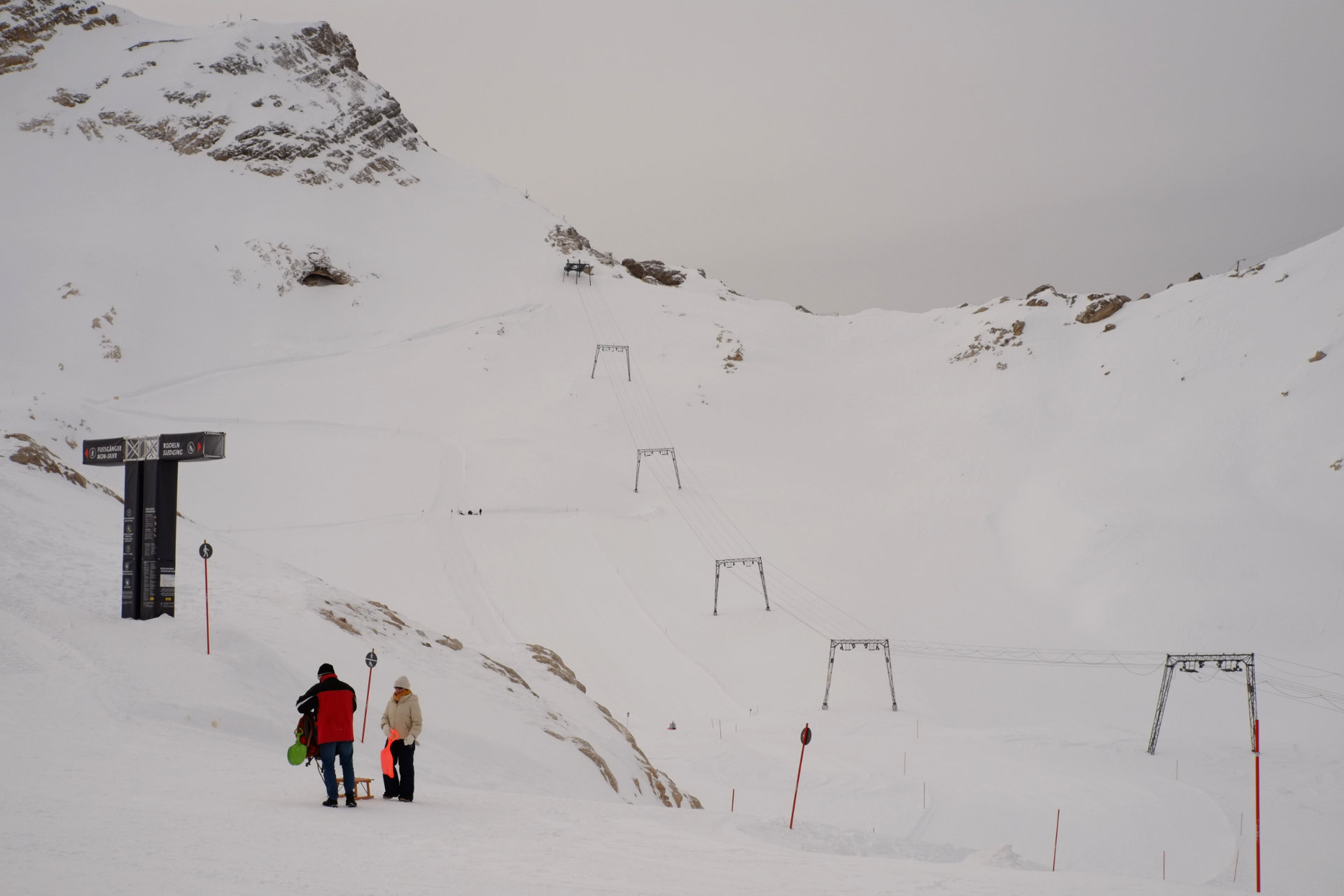 A photo of a big skiing slope going up to the mountain ridge and two people with sleds staying in front of the slope.