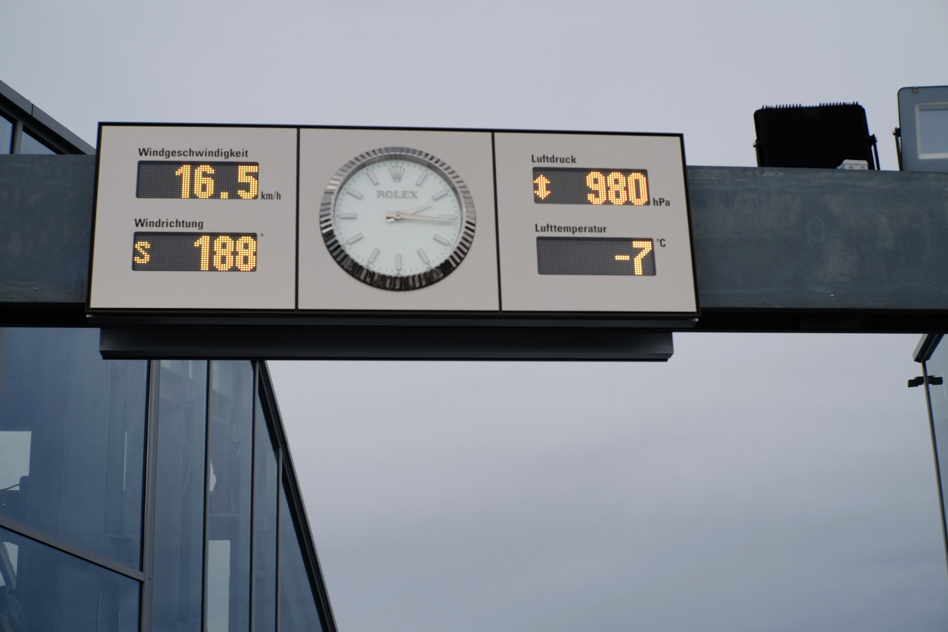 A photo of a clock panel bolted onto a dark metal bar. The panel is also showing the temperature, wind direction, wind speed, and air pressure.