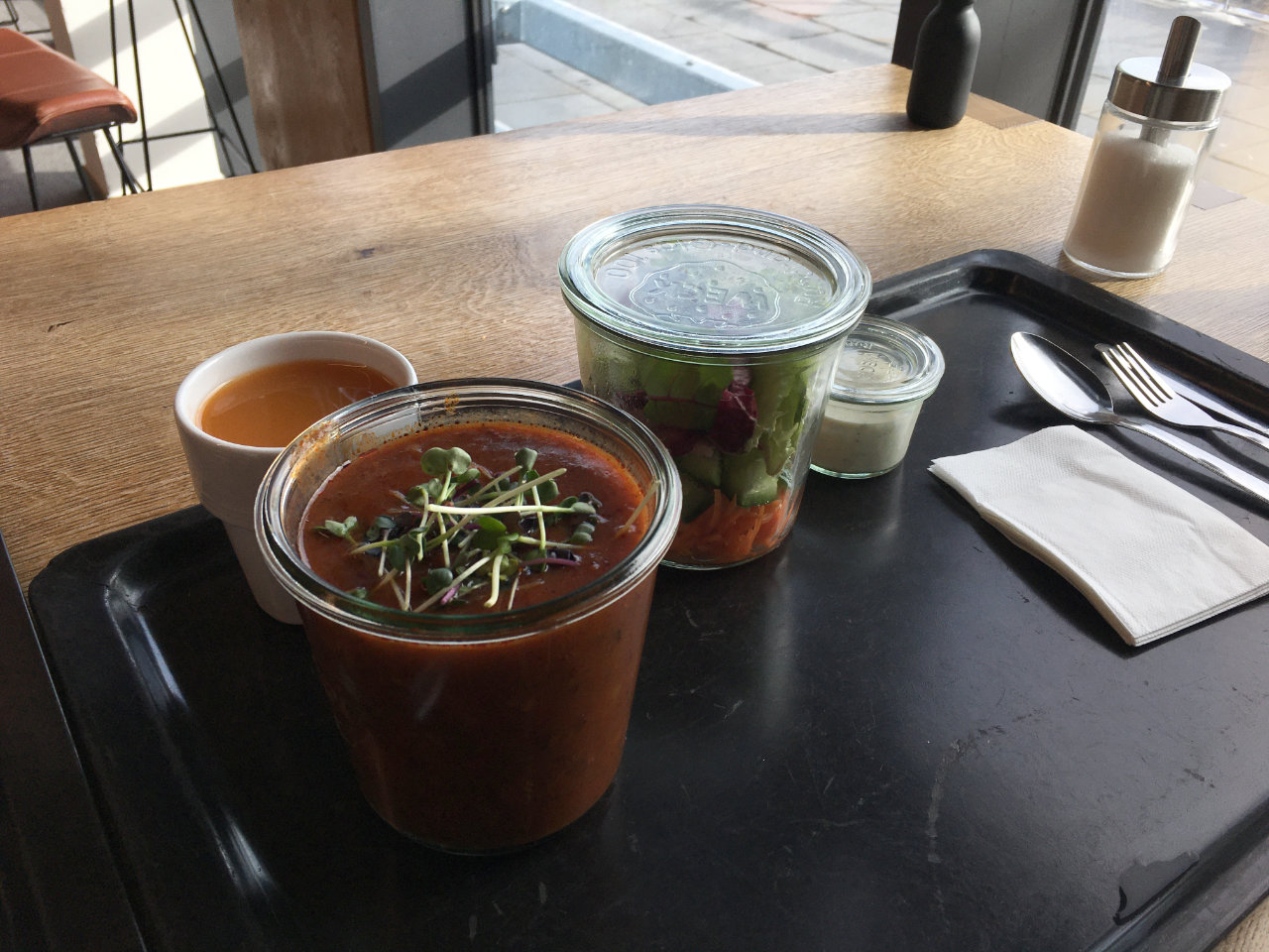 A photo of a canteen tray with two glass jars on it, one with a red-brown soup and another one with salad, one small glass with a salad sauce, and one glass with juice.