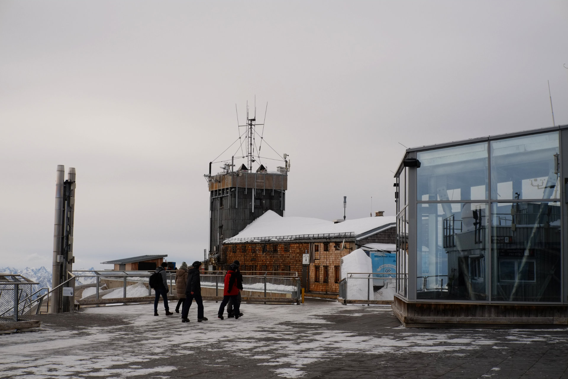 A photo of the observation deck with a group of people moving across it. To the right is a glass structure, in the back there is a dark square tower, the ground is lightly covered with snow.