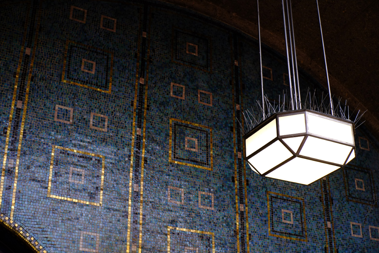 A photo of a blue and golden mosaic wall at the north entrance of the Alter Elbtunnel. An old lamp in front of it is covered with anti-pigeon metal spikes.