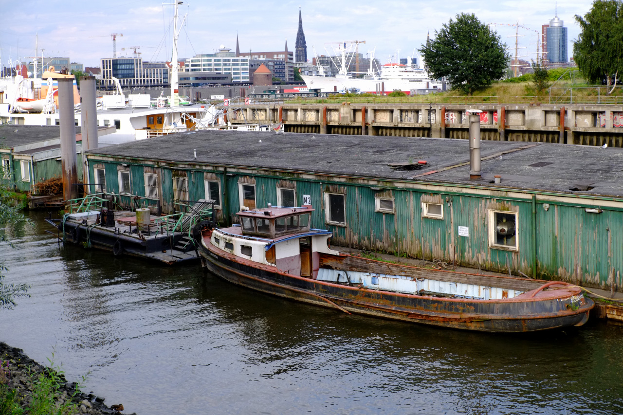 A photo of a shipyard featuring a long green wooden building on water with a rusty boat docket to it.