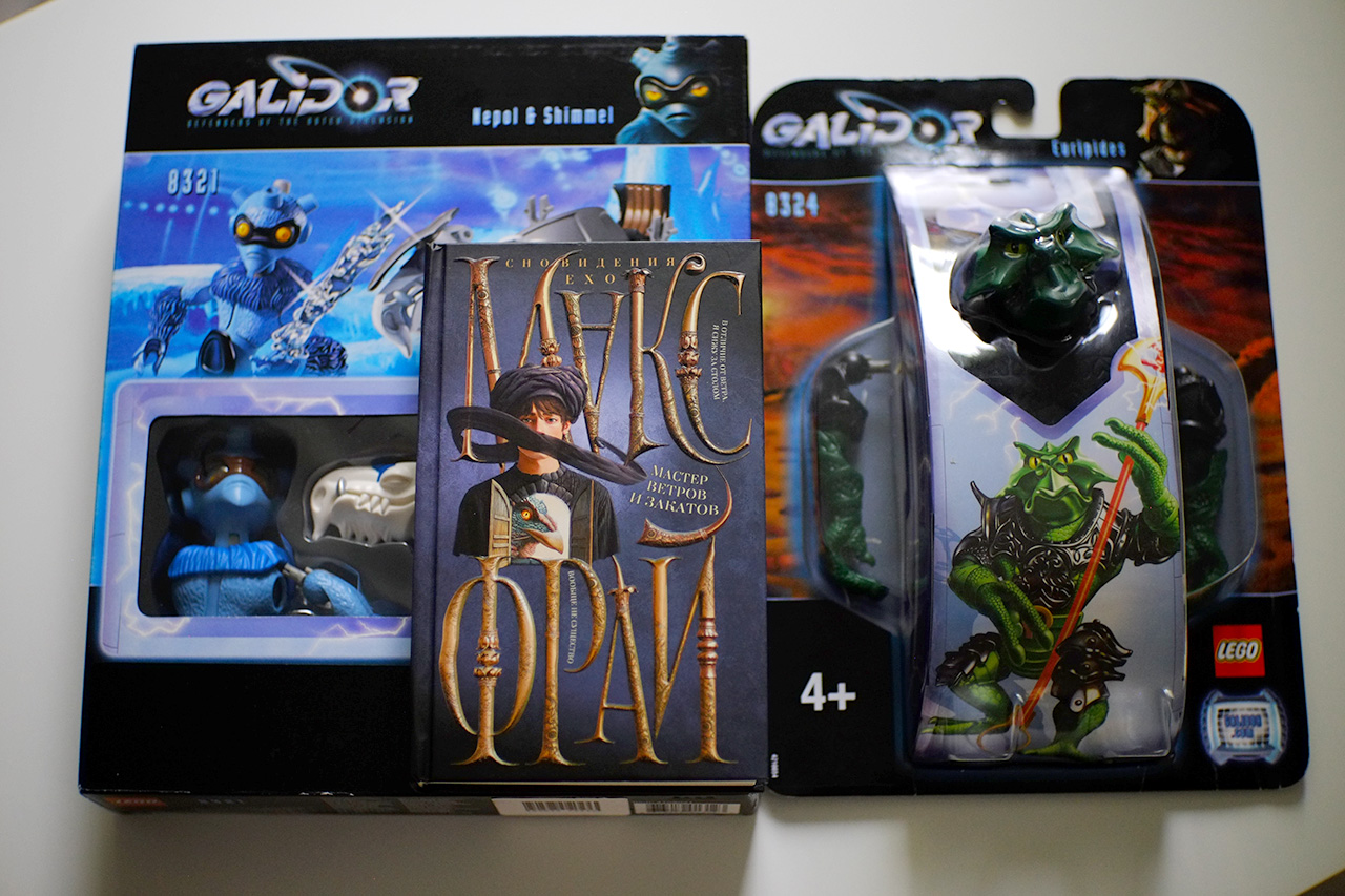 A photo of three objects placed on a white tabletop: a box of LEGO Galidor with a blue and white antropomorphic character with big eye protecting gear and a blue and white furry horse-like creature; a box of LEGO Galidor with a green antropomorphic character in black armor holding a long dark red staff; a book by Max Frei from the "Dreams of Echo" series.