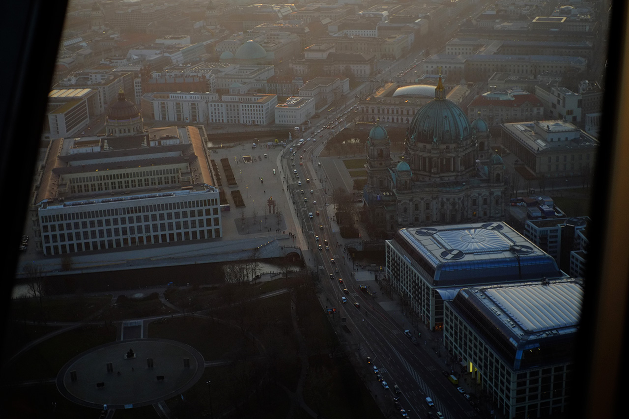 A photo of the view at Berlin from 210 meter height, featuring a round square with a monument in the center, a wide road with moderate traffic, and two museum buildings: one of them has a big green copper dome with four smaller green copper domes.