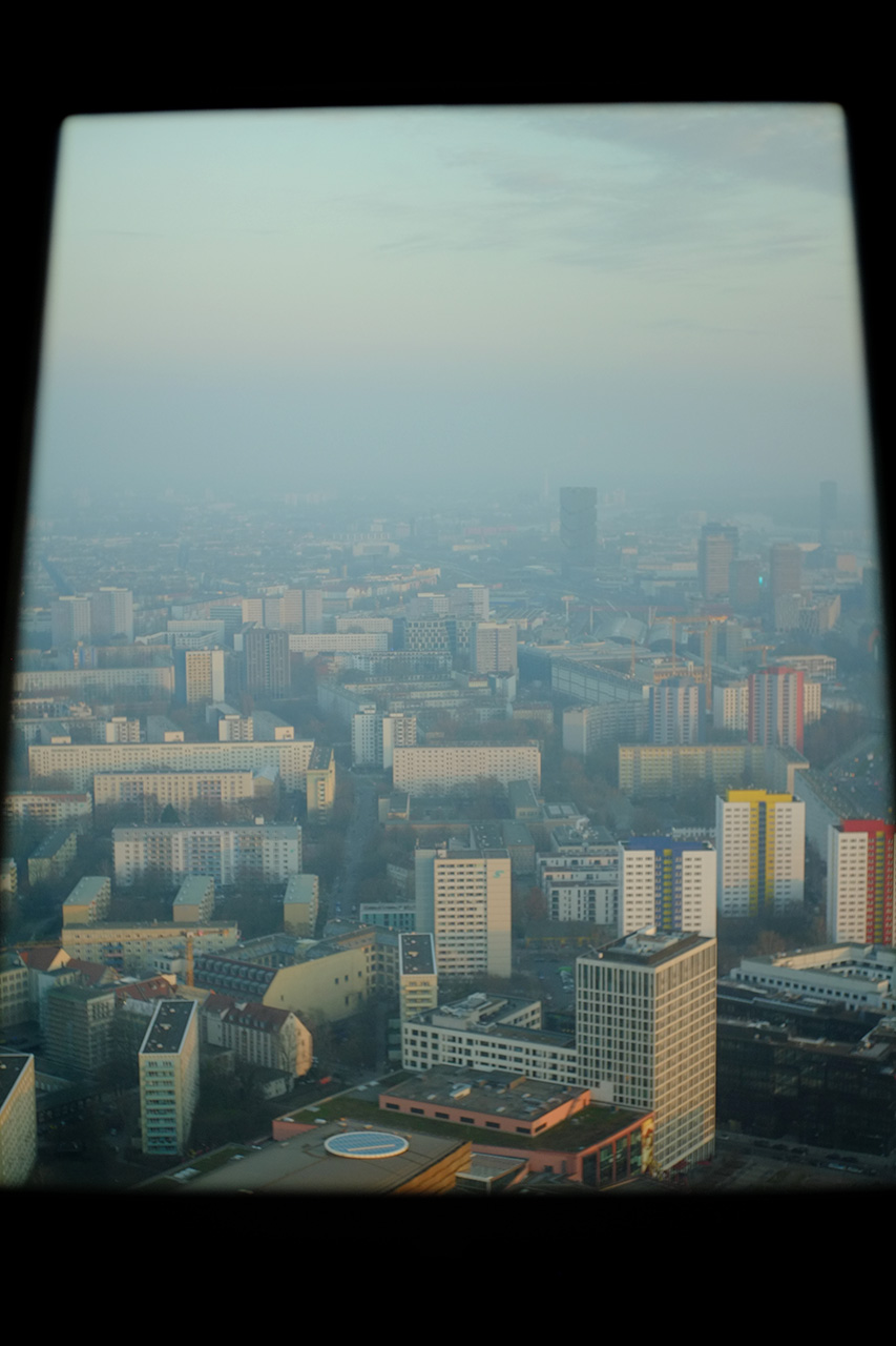 A photo of the view at Berlin from 210 meter height. The view is framed by dark window frames. There are a lot of high-rise residential buildings with a wide prospect road visible. The city is covered in smog and it's almost sunset time.