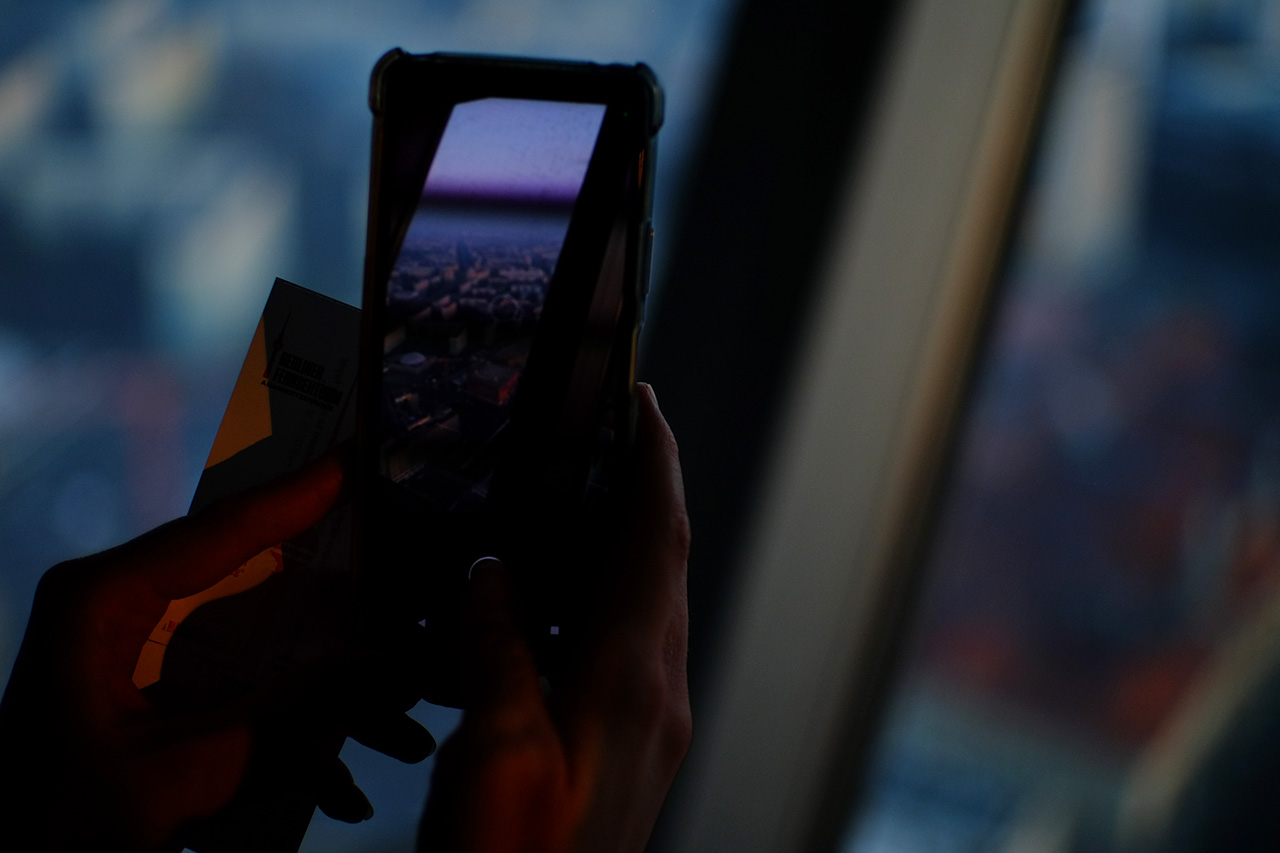 A photo of two hands illuminated by sunset light holding a smartphone while making a photo of Berlin from above.