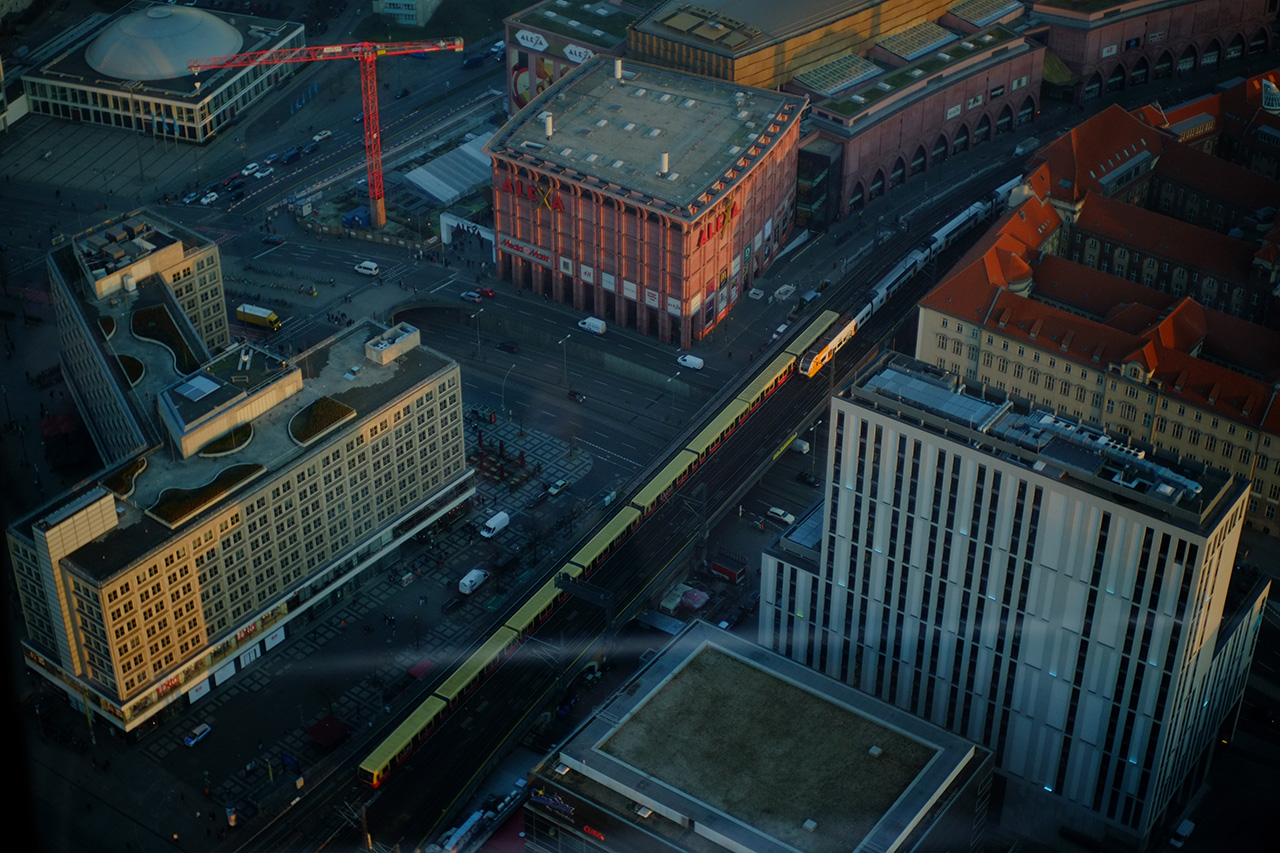 A photo of the view at Berlin from 210 meter height, featuring buildings near Alexanderplatz with a railway track going between the buildings. Two trains are on the railway track going in the opposite directions.