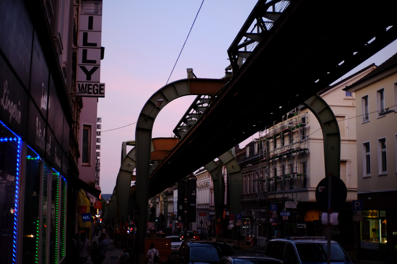 A photo of a narrow street in Wuppertal with suspension railway tracks going above it. The sky in the background is light pink from sunset.