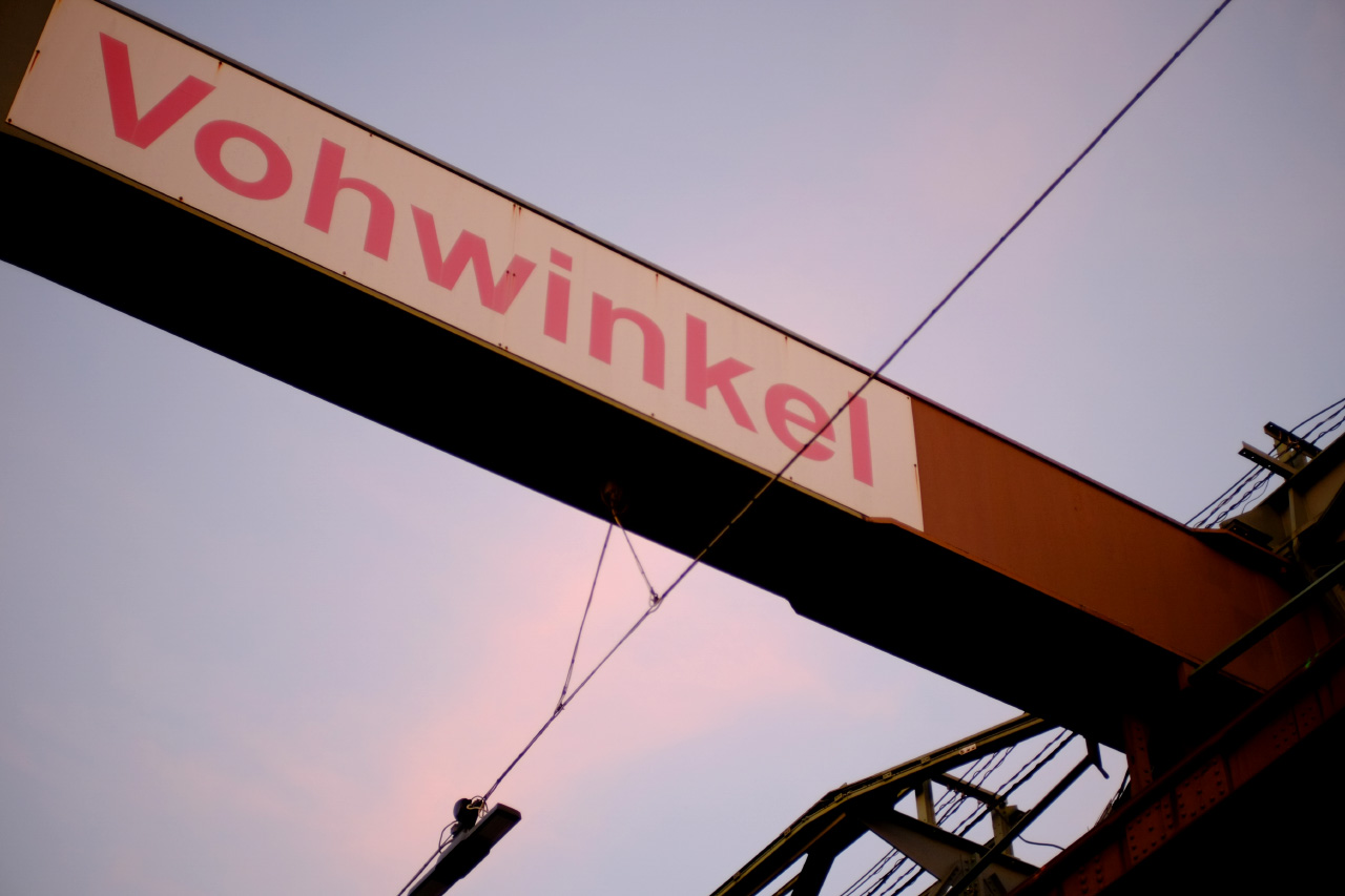 A photo of a suspension railway station sign saying "Vohwinkel" in red with pink and lilac sunset sky in the background.
