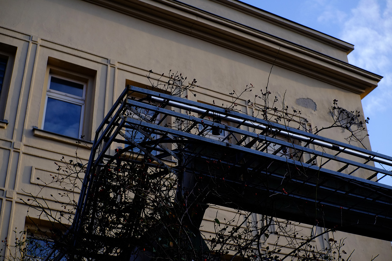 A photo of a corner of a light-gray building with several tall windows and a black metal ladder in front of it.