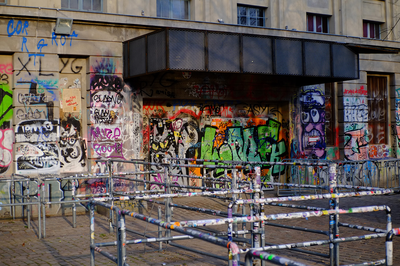 A photo of an entrance to a light-gray building: the wall with the entrance doors are all covered in graffiti and metal fencing in front of it is all covered in stickers.