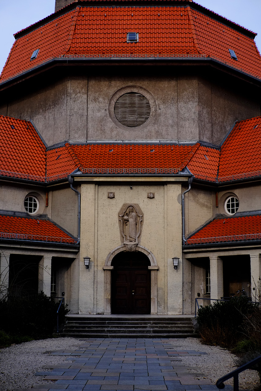 A photo of the inner yard of an old looking building with a big red dome and two two-storey building wings surrounding the yard. The central entrance has a big dark wood door and a bas-relief sculpture above it of a person in a long robe with their hand above an urn.
