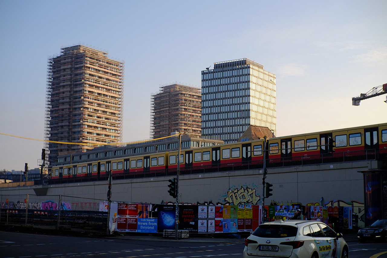 A photo of an intersection of a railway track and a road: the railway track is above ground and a red and yellow train is moving on it; the road goes into a tunnel under the railroad track; concrete foundation of the railway track is all covered in graffiti.