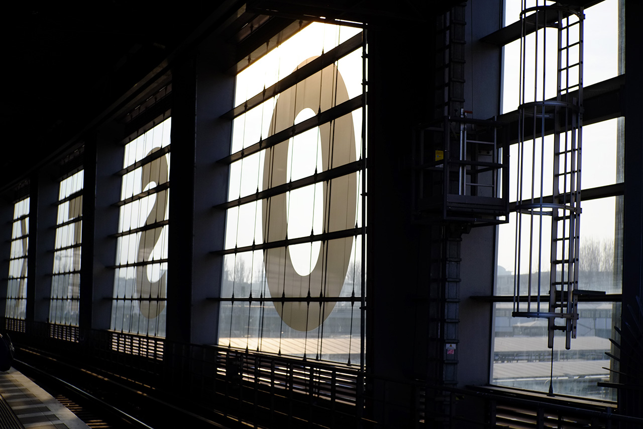 A photo of one of the sides of the Ostkreuz train station featuring a glass wall with big metal bars and big white letters O, S, T, K printed on the glass wall sections.