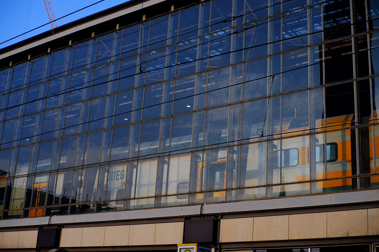 A photo of a train station from the outside. The station is build above ground and has glass walls. Behind the glass wall a white and yellow train can be seen.