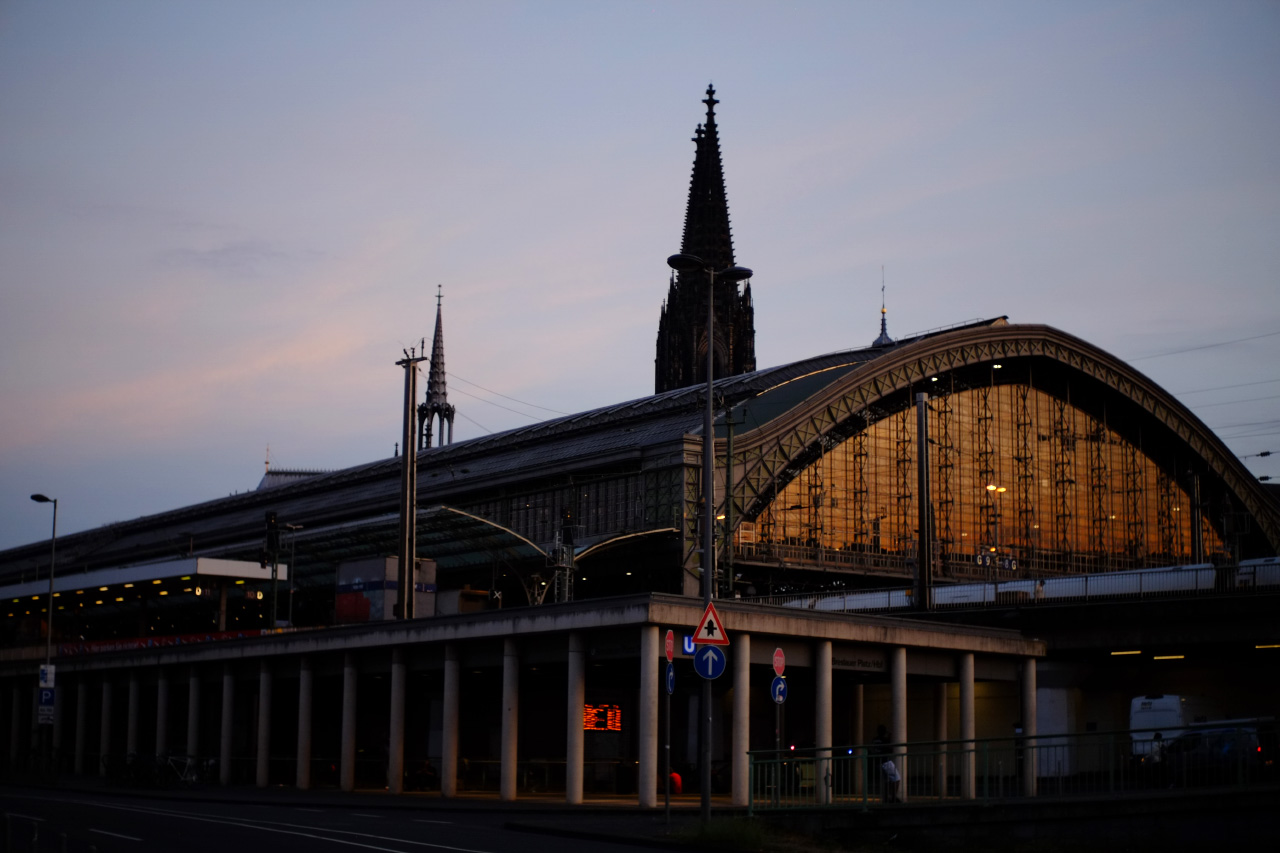 A photo of the main building of the Köln central railway station with it's big windows reflecting the sunset sky. Dark spires of an old cathedral are visible behind the building.