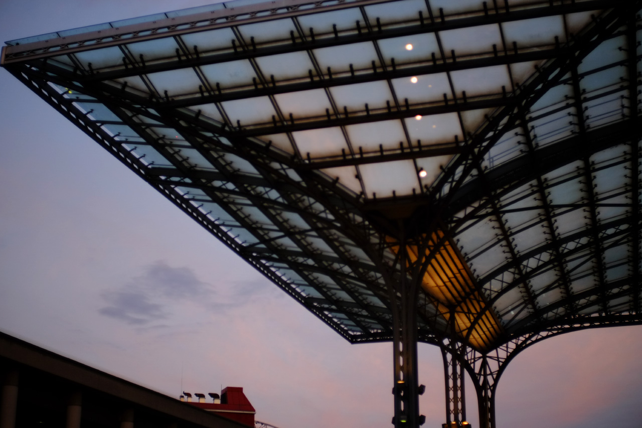 A photo of an arched glass overhang in front of the main railway station building with lilac sunset sky in the background.
