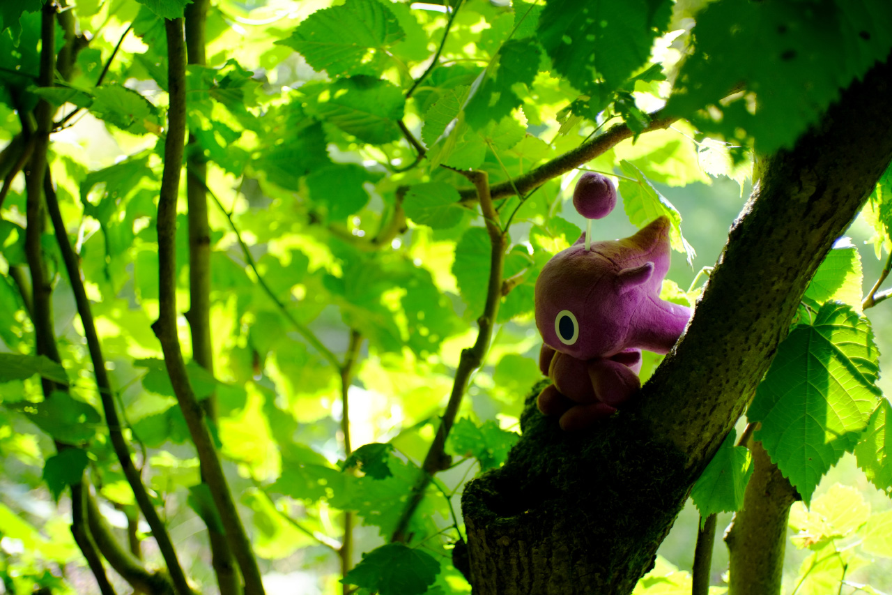 A photo of a violet plush toy of a small creature sitting on a small tree with green leaves in the background.