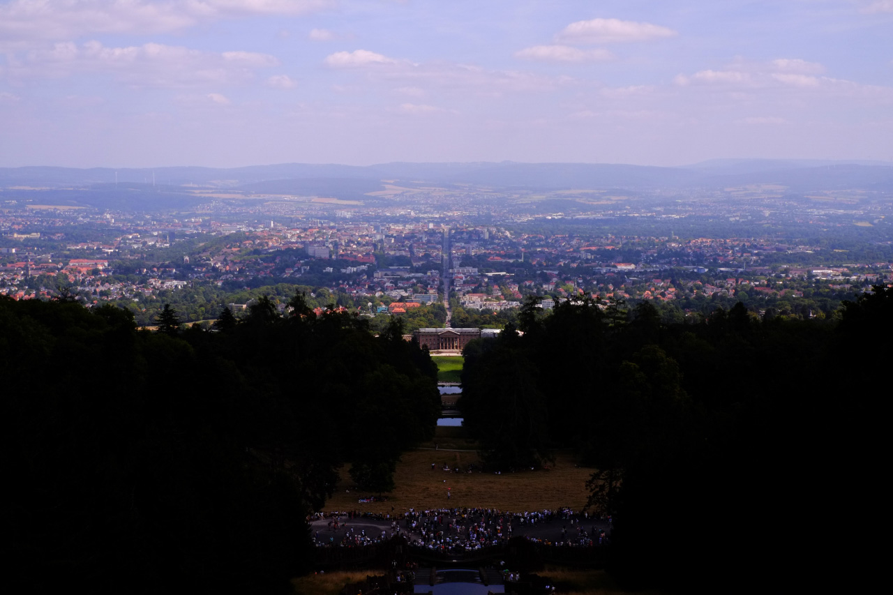 An overview photo of Kassel from the Hercules pedestal, featuring a long stretch of land in the center of a forest that goes down the hill to a big old castle building and a big city visible behind the building.