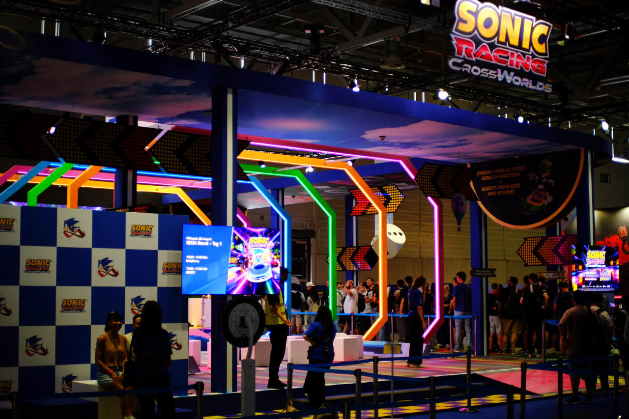 A photo of the Sonic Racing CrossWorlds booth at gamescom 2025, featuring a podium for taking photos, a big space with colorful neon arches and big white cubes to sit on, a ceiling tile with blue sky and white clouds printed on it, and a big game logo hanging above the booth.