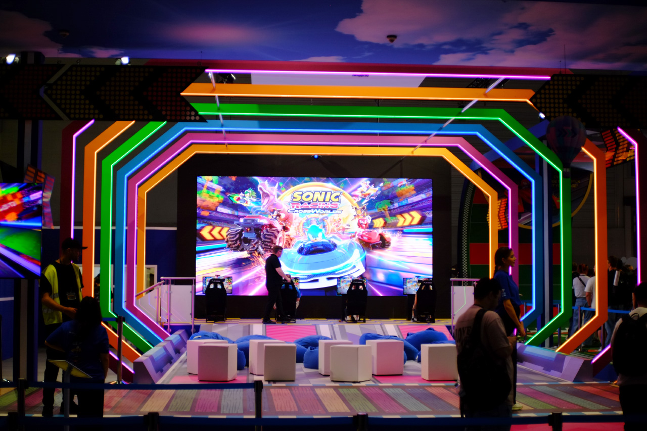 A photo of the big open booth space with colorful neon arches, a big screen in the back, four gamer chairs with computers set up in front of them, blue bean bags in the shape of Sonic the Hedgehog head, and white cubes to sit on.