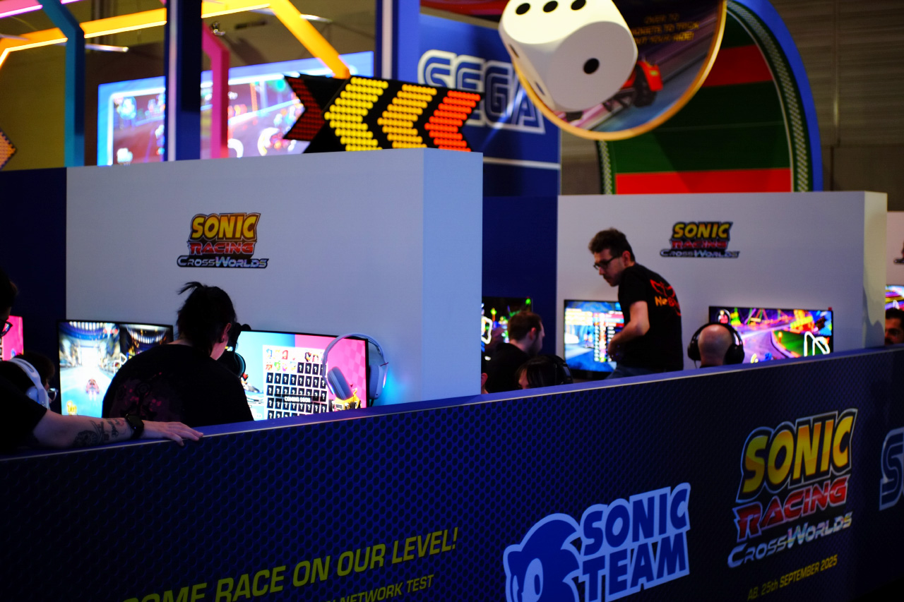 A photo of the Sonic Racing CrossWorlds booth segment at gamescom 2025 with screens mounted onto black and blue space dividers. There are two booth workers helping visitors with the game demo.