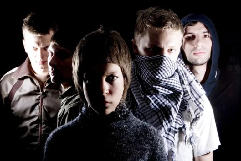 A photo of five band members standing in a V-formation in the dark and their bodies are being illuminated by one strong direct light.