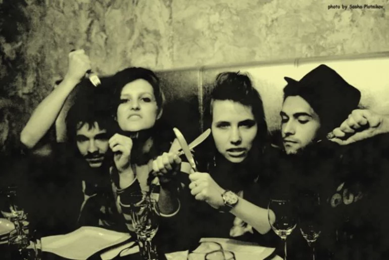 A photo of two young men and two young women sitting at the restaurant table and demonstrating the kitchen utensils in their hands with threatening looks at the camera.