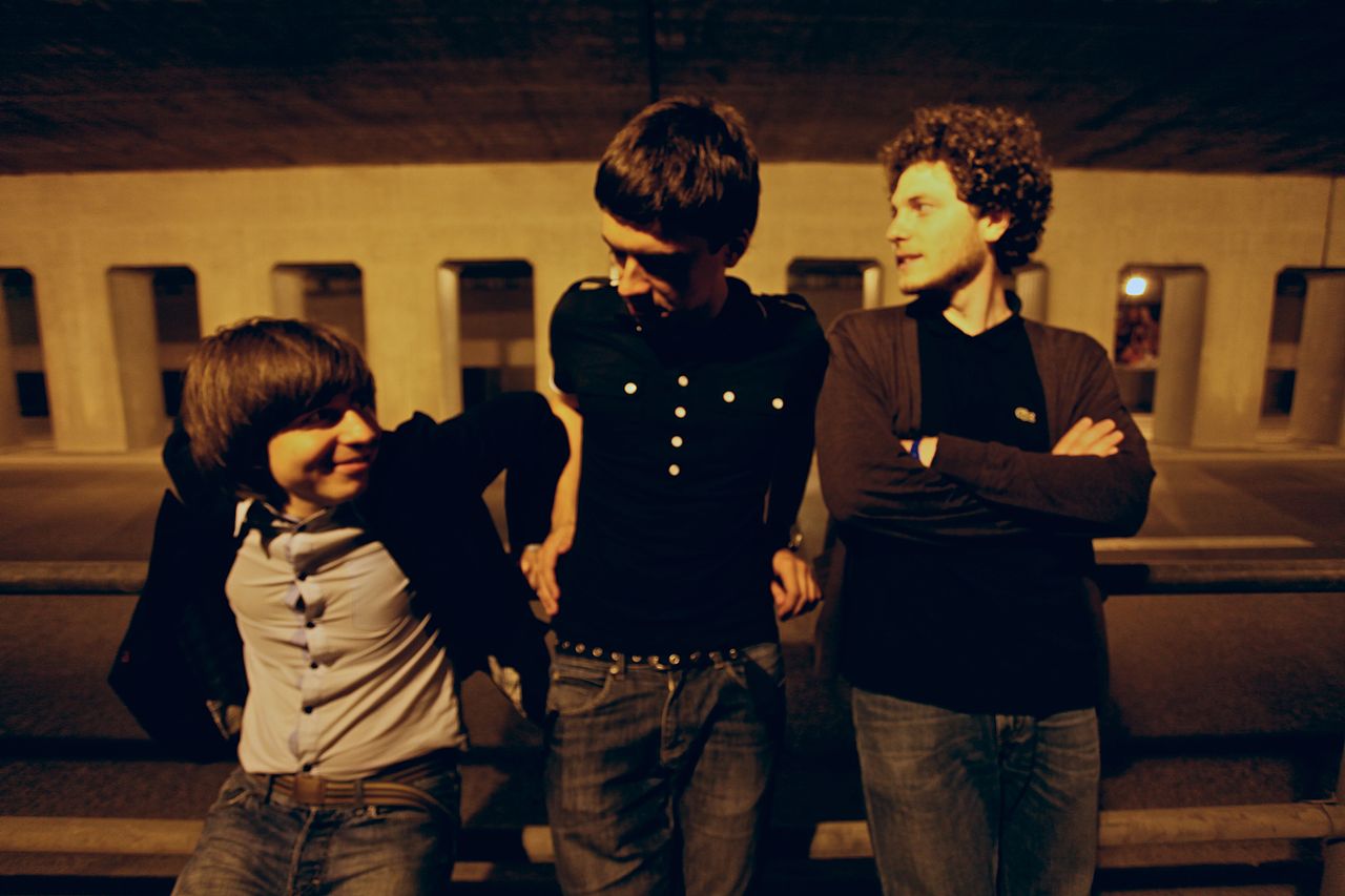 A night photo of three young men posing in front of a concrete structure with arches. They are avoiding looking at the camera.