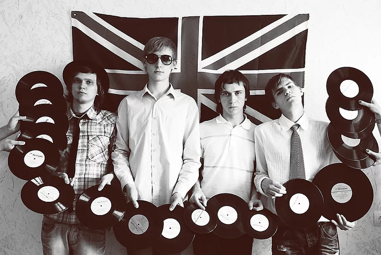 A black-white photo of four young men posing in front of the Union Jack flag and holding a lot of vinyl records in their hands.