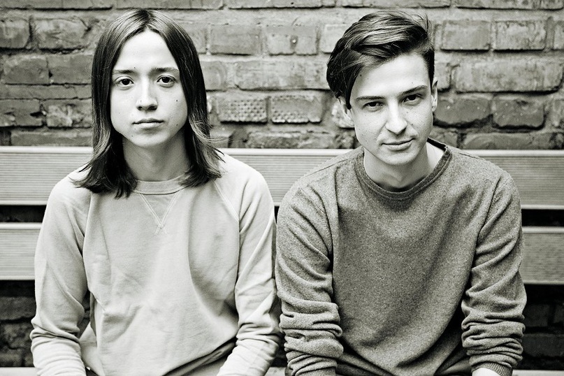 A black-white photo of two young men sitting on a bench in front of a brick wall and looking at the camera. Both are wearing sweatshirts.