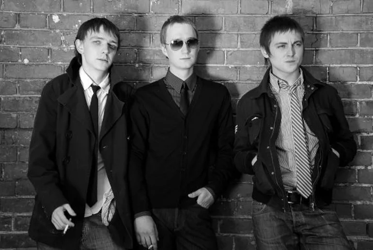 A black-white photo of three young men in front of a brick wall dressed in jackets, shirts, and ties. Two of them are holding cigarettes between their fingers.