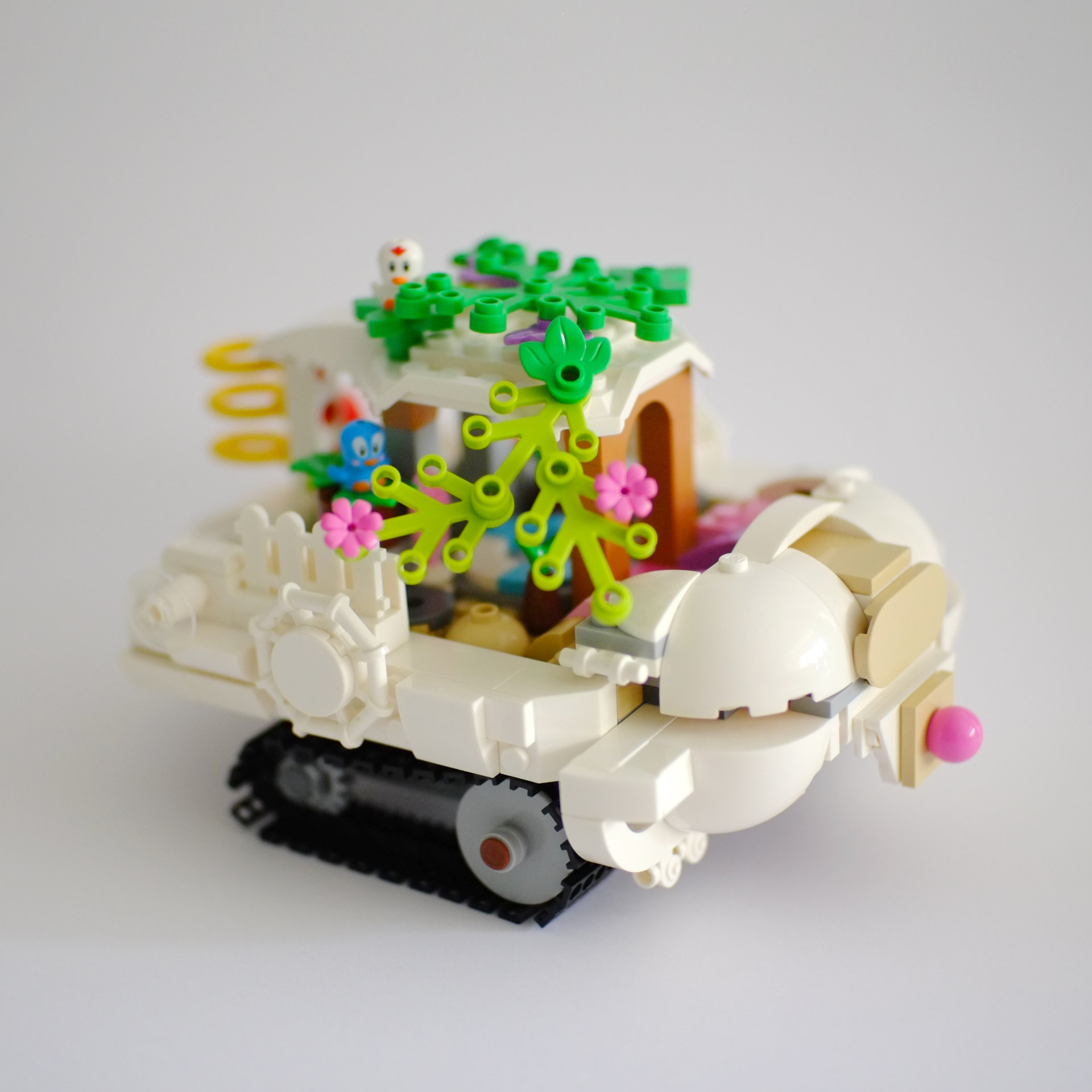 A photo of the right side of a lego build that is a white tourist boat on tank tracks. The roof of the boat is covered with foliage with birds sitting on top.